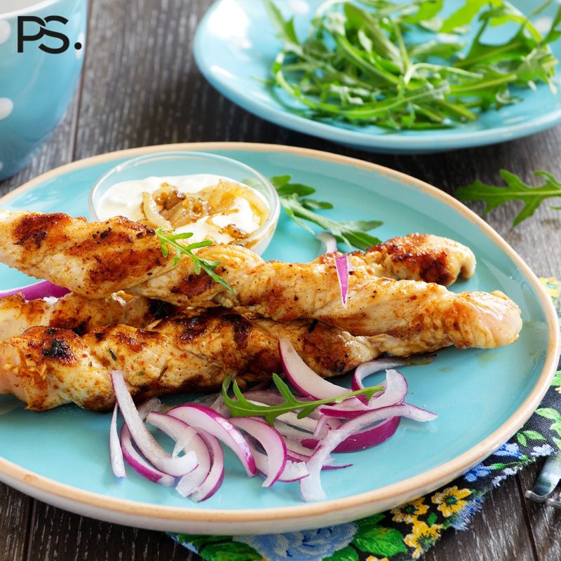 Gegrillte Hähnchenspieße mit roten Zwiebeln, Rucola und Dip werden auf einem blauen Teller angerichtet.